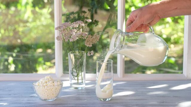 Pouring fresh milk into a transparent glass from jug on the windowsill by the window of the house on a summer day near the garden, close up