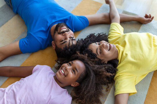 Portrait of happy family posing on floor at home, laying on carpet and bonding, cheerfully smiling, view above. Positive father, mother and daughter cuddling, top view