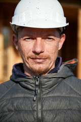 Close up portrait of male builder building wooden frame house. Man standing on construction site in white safety helmet with dirty face after painting.