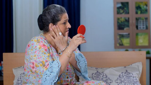 A frustrated upset Indian lady touching her face while looking in a small mirror - dry sensitive skin  aging. A worried elderly woman depressed about facial wrinkles and dark circles at home - an I...