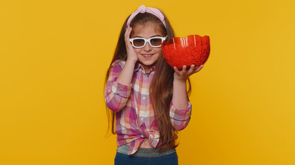 Little toddler children girl in shirt eating popcorn, watching comedy movie film wearing 3D glasses, enjoying cinema snacks. Young preschool kid in glasses isolated alone on yellow studio background - obrazy, fototapety, plakaty