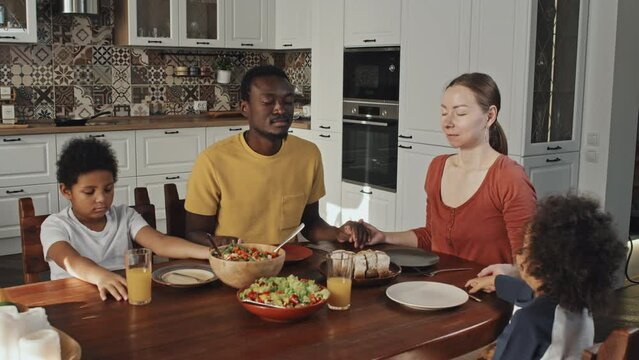 Medium of young Caucasian woman, African American man and their two adorable sons sitting at dining table, praying before lunch