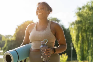 Young African American woman walking with exercise mat and bottle of water through the park in a summer day © gpointstudio