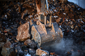 The bucket of a construction excavator scoops up destroyed concrete and bricks and large piles.