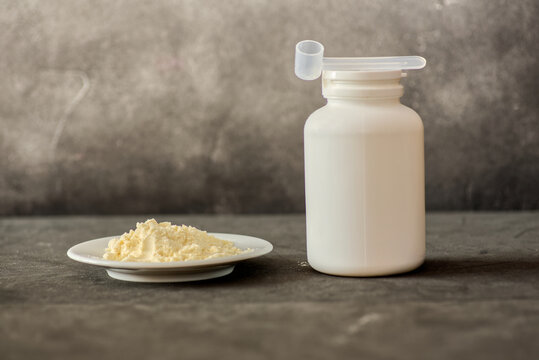 Heap of bovine colostrum powder on plate and white plastic container with measuring scoop on dark background. Colostrum health benefits concept