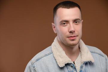 A young man in a casual jacket on a brown background looks at the camera and smiles slightly