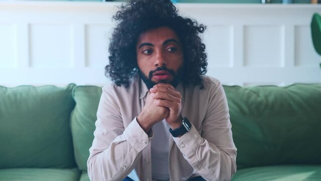 Young perplexed Arabian man nervous thinking about problems and troubles that have arisen in career or business trying to find solution and fussy touching face with hands sits on sofa in own house