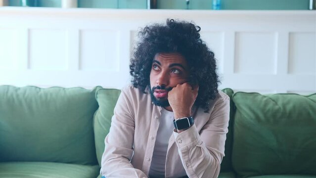 Young bored curly middle eastern man in casual clothes feels longing from idleness props head with hands when watching uninteresting movie or waiting for long awaited call sits on sofa in apartment.