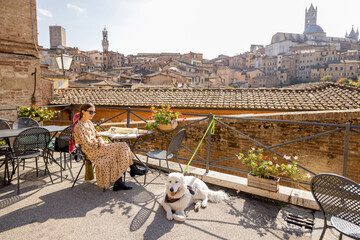 Young woman having lunch with pizza and wine, sitting with dog at outdoor restaurant with beautiful view on the old town of Siena. Concept of italian cuisine and travel - obrazy, fototapety, plakaty