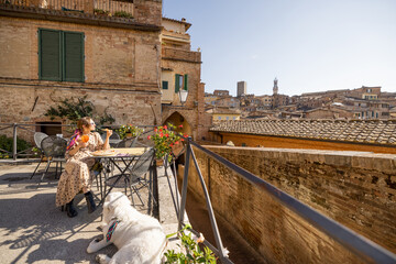 Young woman having lunch with pizza and wine, sitting with dog at outdoor restaurant with beautiful view on the old town of Siena. Concept of italian cuisine and travel - obrazy, fototapety, plakaty