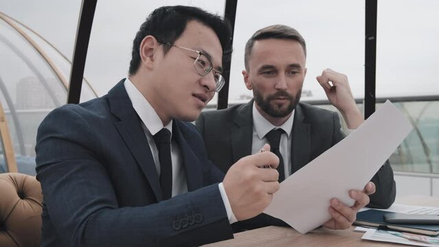 Low angle waist-up of two Caucasian and Asian male top managers in formal suits sitting at table in outdoor cafe at daytime, working with documents, having conversation