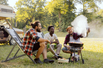 A group of friends spend time together in nature. The middle-aged men prepare a barbecue near the RV. Bearded boy checks the grill