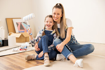 Two female sisters are sitting on floor in room that is under renovation. One girl 28 years old has charming smile long hair, green eyes. Second girl, aged 8 is wearing beige trainers checkered shirt.