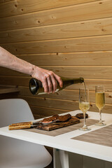 Man pouring white wine from a bottle into a flute glass, cooked and sliced steak meat served on wooden cutting board with cutlery on the table