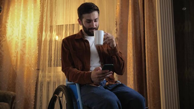 Young man in a wheelchair drinks tea and uses his phone for online shopping. An incapacitated guy holds a phone and a white cup sitting at home.