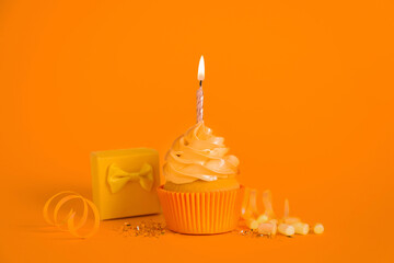 Delicious birthday cupcake with burning candle, marshmallows and gift box on orange background