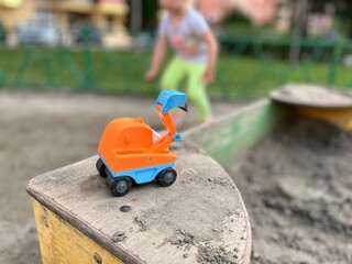 Bright plastic toy excavator in the sandbox on a playground. A child runs and plays near their toy excavator on a playground in summertime