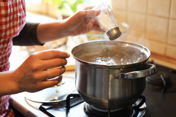 Woman adding salt to boiling water in pot on stove indoors, closeup