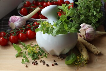 Mortar with fresh herbs near garlic, horseradish roots, black peppercorns and cherry tomatoes on wooden table, closeup