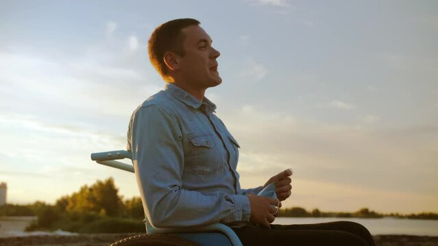 young man in a wheelchair takes off his medical mask in the street and rejoices. Disabled man takes off his mask and smiles for the camera.Disabled man against the backdrop of a sunset.
