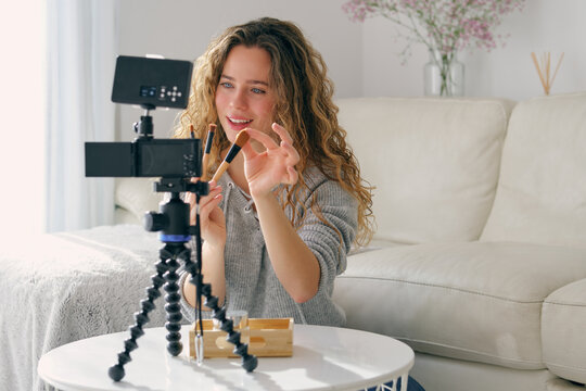 Young female vlogger with long wavy hair showing set of brushes for facial makeup on camera in living room at home