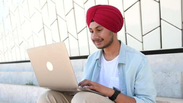 Indian freelancer man in traditional turban sitting outdoors with laptop, developer working on the distance, young guy enjoys remote work, male student studying online, e-learning concept