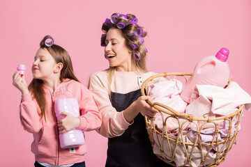 Smiling mother and daughter pose against pink background with a basket of laundry clothes. The sweet little girl sniffs the cap the beautiful scent of freshness of clean clothes floral washing liquid.