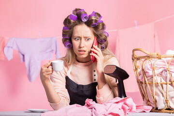 Terrified woman answers phone and learns shocking facts, worried expression on face. Girl has rollers on head next to steamer for ironing clothes morning in laundry room over coffee and conversation.