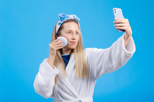 Influencer girl takes photos of herself and records a video during the morning facial care. Young woman in a bathrobe on a blue colored studio background.