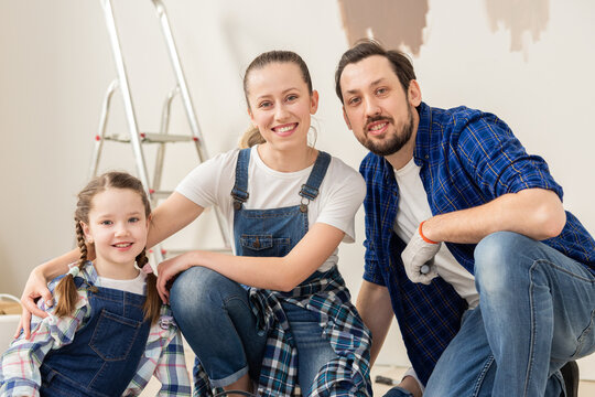 A happy family, dressed in denim style clothes, sits in the middle of their newly renovated apartment. In the background there is a ladder for painting the wall.