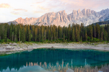 Stunning view of Carezza Lake (Lago di Carezza) with its emerald green waters, beautiful trees and mountains in the distance during a dramatic sunset.