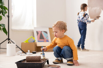 A very cute 2-year-old child, dressed in an orange blouse, jeans and sneakers, is sitting in the middle of the room where repairs are taking place. The boy holds a paintbrush in his right hand.