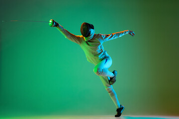 Energetic young man, fencer with smallsword practicing fencing isolated on green background in neon...