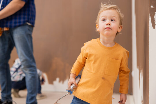 Small boy dressed in an orange blouse holds a paint roller in his hands smeared with paint. The child is standing in the room where repairs are taking place and is thinking about something.