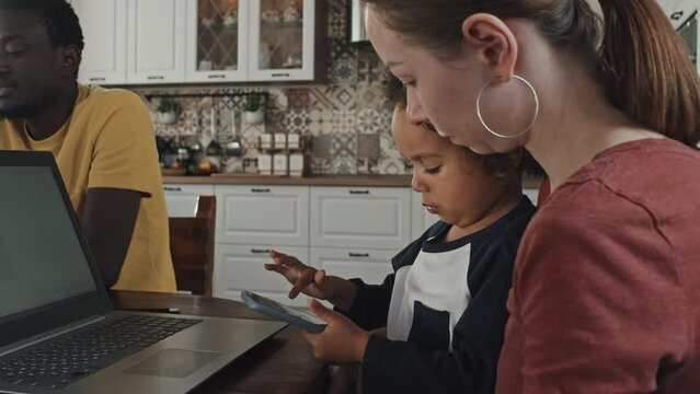 Low Angle Side View Of Young Caucasian Woman Sitting At Table With Little Black Son On Knees, Boy Using Smartphone