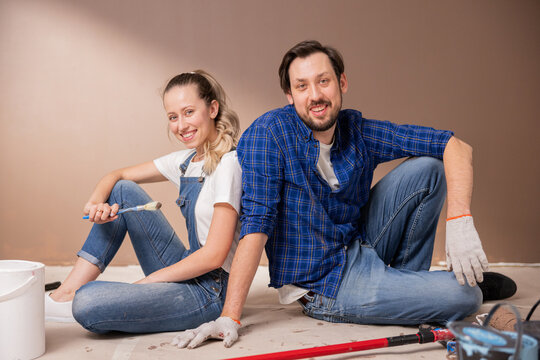 a blonde woman and her husband, a brunette with a mustache and a beard, sit back to back on the floor in a room where renovations have begun. The girl is holding a paint brush.