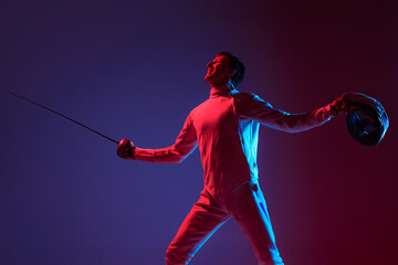 Portrait of young tall male fencer in fencing costume and mask holding smallsword isolated on purple background in neon. Sport, emotions, energy, skills