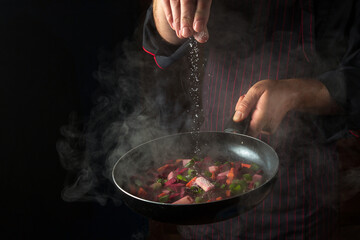 An experienced chef salts vegetables in a hot frying pan. Menu concept for hotel on black background