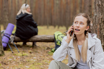 Woman with blonde hair tied in braids sits on the ground in the forest and talks on the phone, resting while trekking, camping, hiking.