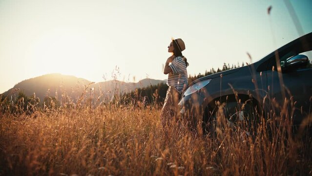 Cinematic footage of a young woman standing leaning on the hood of her car and enjoying the sunset during her solo road trip.
