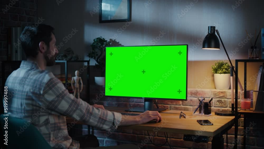 Poster Young Handsome Man Working from Home on Desktop Computer with Green Screen Mock Up Display. Creative Male Checking Social Media, Browsing Internet. Loft Living Room in the Evening. Zoom Out Shot.