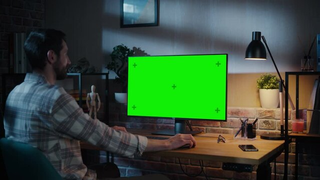 Young Handsome Man Working from Home on Desktop Computer with Green Screen Mock Up Display. Creative Male Checking Social Media, Browsing Internet. Loft Living Room in the Evening. Zoom Out Shot.