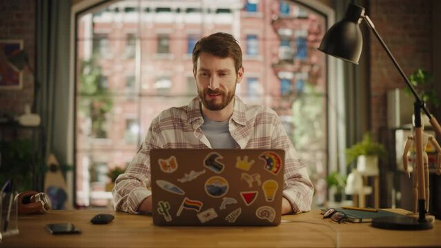 Handsome Bearded Young Adult Man Having an Online Conversation on a Laptop Computer in Creative Office Environment. Happy Specialist Browsing Internet and Social Media Platforms.