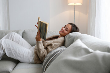 Young beautiful woman wearing knitted cardigan reading a book at home. Brunette female in a beige...