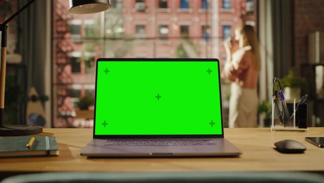 Laptop Computer Display with Mock Up Green Screen on a Table in Living Room. Chroma Key Monitor at Home or Creative Loft Office with Female Drinking from a Mug and Looking Out of Window in Background.