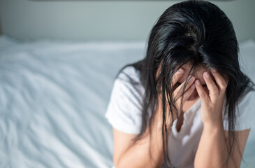Fototapeta premium Asian woman in white shirt sitting in bed holding her head expressing stress, disappointment, depression, migraines, health issues, health care and stress management concept.