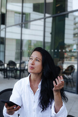 Mature beautiful woman sitting at a street cafeteria with smartphone and smoking cigarette