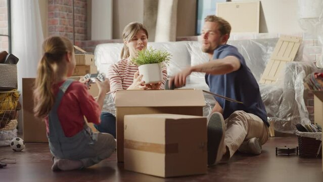 Happy Homeowners Moving In: Lovely Couple Sitting on the Floor of Cozy Apartment Unpacking Cardboard Boxes, Little Daughter Joins them. Cheerful Day, Happiness, Sweet Home for Young Family Having Fun