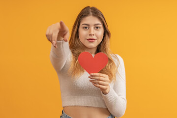 A young girl with long hair holds a heart made of paper and points to the camera. It's like choosing a partner. Dating site concept.