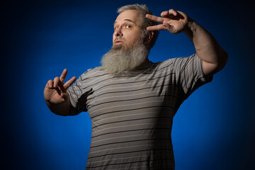 A gray-haired, bearded, disheveled adult man in a striped shirt is dancing, waving his arms wide. The man looks into the camera. Dance concept for old people.